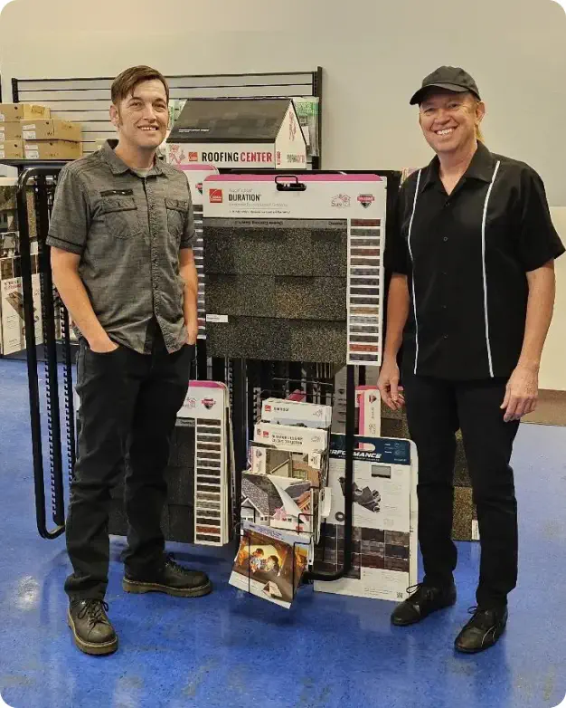 Two smiling men standing in a home improvement store aisle beside a display of roofing materials, one is younger wearing a gray shirt, the other older in a black shirt and cap.